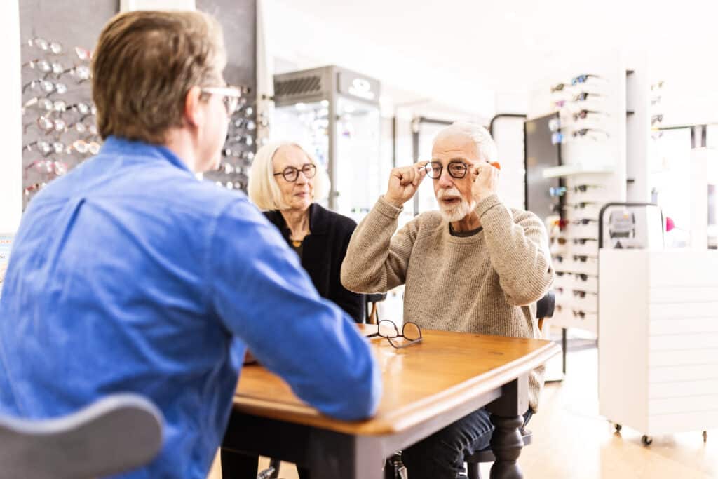 Zwei Personen werden in einem Brillenfachgeschäft von einem Optiker beraten und probieren ein Brillengestell an